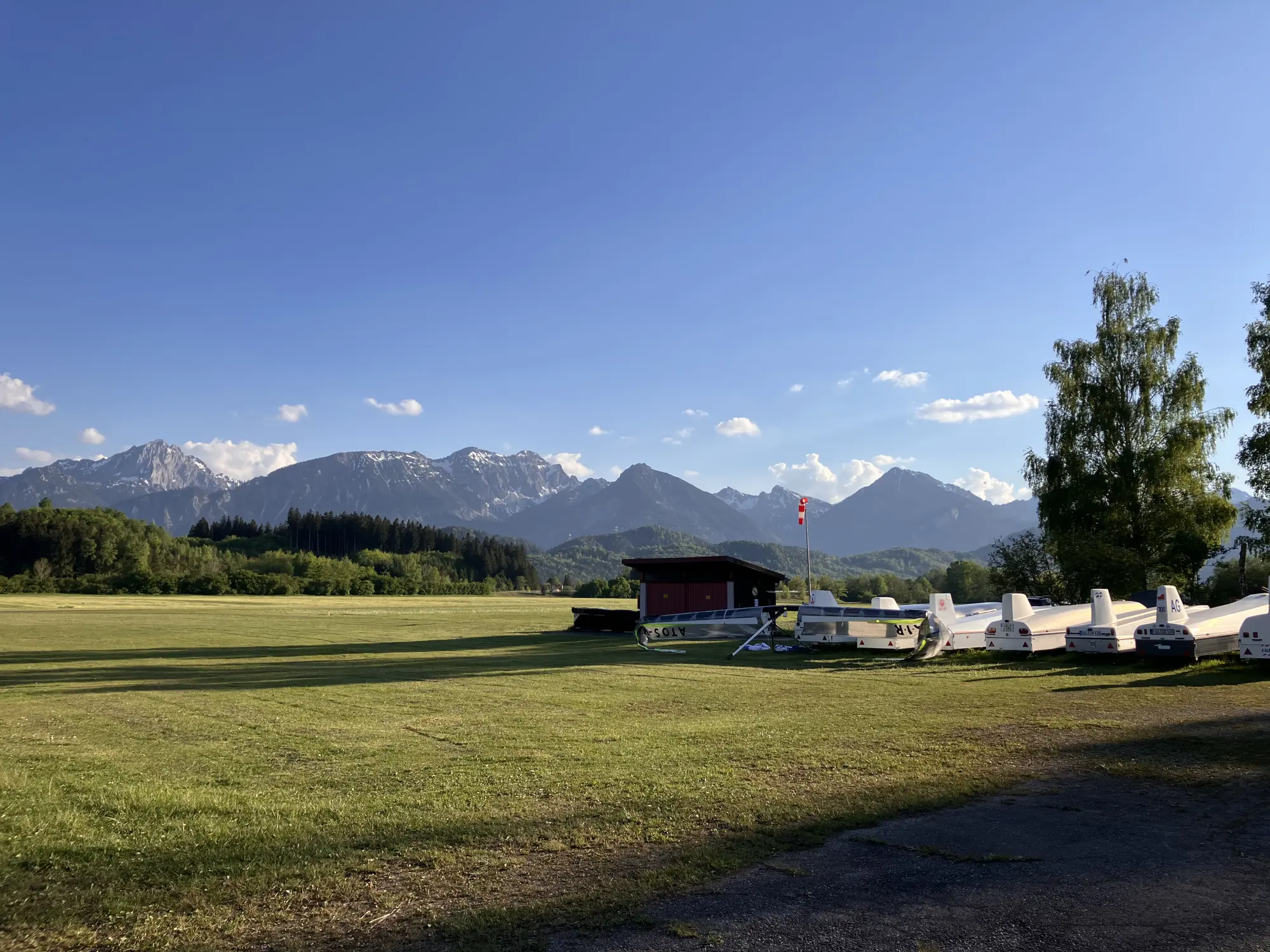 Segelfluggelände mit Anhängern und Windsack vor den Alpen